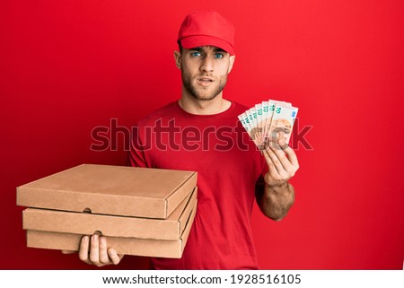 Young caucasian man holding take away food and 10 euros banknotes in shock face, looking skeptical and sarcastic, surprised with open mouth 