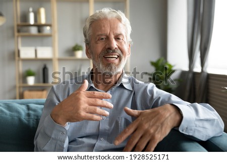 Mature man sitting on couch looking at camera having distant conversation with grown up children using web camera online application. Concept of modern technologies and older generation easy usage Royalty-Free Stock Photo #1928525171