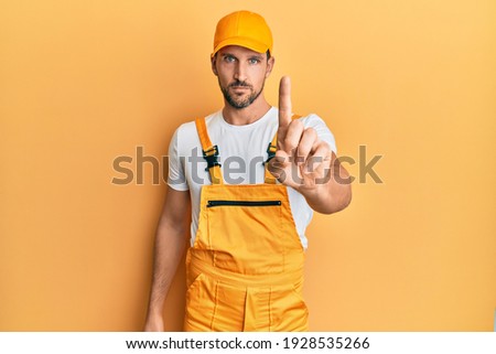 Young handsome man wearing handyman uniform over yellow background pointing with finger up and angry expression, showing no gesture 