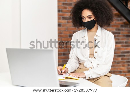 African American female student or businesswoman with curly hair wearing black protective face mask sitting in the coffee shop or library, doing research, looking at laptop, taking notes to notebook
