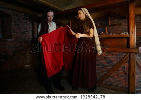 Girls in historical costumes. Medieval clothing. Two maids in vintage dresses are buying red cloth. Reconstruction of a past life. Tavern waitresses. Wooden bar counter in a cafe. Two models talking Royalty-Free Stock Photo #1928542178