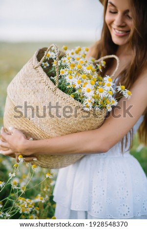 Cute young woman looks at daisies in a basket in sunny weather Beautiful white dress on her body and a hat on her head Brunette with long hair