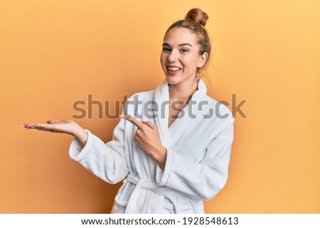 Young blonde woman wearing bathrobe amazed and smiling to the camera while presenting with hand and pointing with finger. 