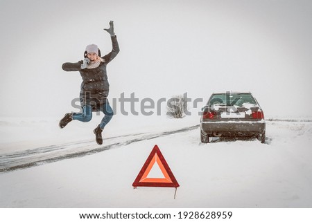 Enraged young woman is talking on a mobile phone, jumping in anger near a warning road sign-a triangle against the background of a car on a snow-covered road. Concept of car breakdown