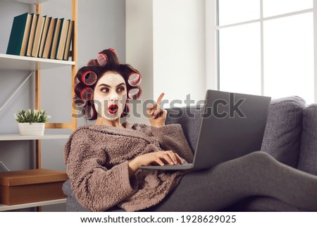 Surprised woman points finger up as she finds amazing idea online. Happy lady in curlers and face mask sitting on sofa, browsing Internet blog on laptop and learning good life hacks and practical tips
