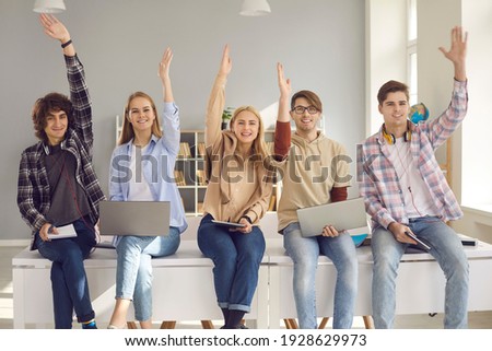 Group of happy smiling active students raising hands sitting on desk with digital devices. Young people agree with good idea or answer questions in lecture class. Studying at school or college concept Royalty-Free Stock Photo #1928629973