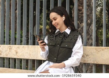 Girl in a green vest smiling on a promenade with palm trees while looking at the mobile