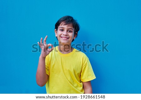 A Portrait of boy in yellow t-shirt making ok gesture with hand, on blue background. Copy space