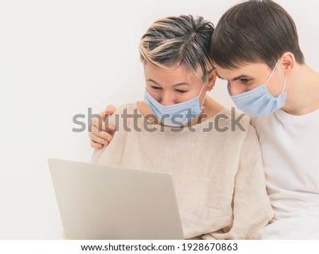 A family in medical masks look at a laptop. The old mother and young son. The concept of self-isolation at home due to the virus pandemic. Spending time during quarantine.