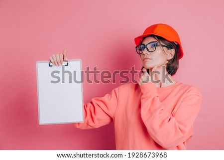 Pretty thoughtful  woman worker builder hold white sign board blank against pink background. Building helmet. 