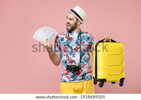 Joyful young traveler tourist man in summer clothes hat hold suitcase fan of cash money in dollar banknotes isolated on pink background. Passenger traveling on weekends. Air flight journey concept