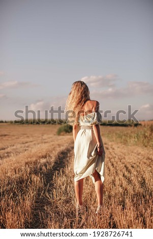 A girl with blond curly hair and in a romantic dress stands in the middle of the field. The wind develops the girl's hair. Lonely girl in nature.