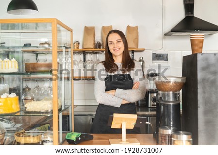Confident female business owner standing at the counter of a restaurant with arms crossed, bakery waitress in apron looks at the camera and smiles