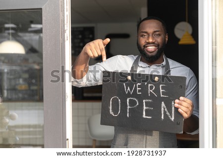 Small business concept. Smiling African-American guy bakery stuff in an apron points finger on an open sign board in his hand. A multiracial cafe owner invites to visit reopened location
