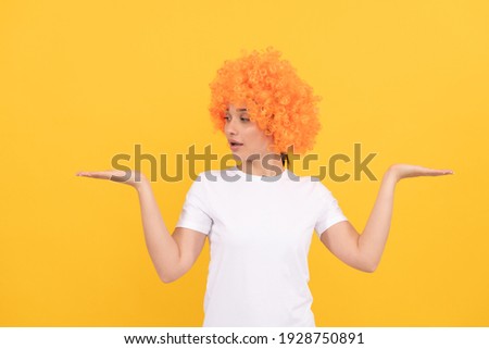 amazed funny girl with fancy look wearing orange hair wig on yellow background presenting product, copy space, choice.