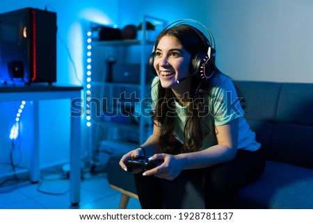 Happy female gamer smiling while playing and winning in a video game with a remote controller. Young woman sitting on the couch in her bedroom with neon led lights Royalty-Free Stock Photo #1928781137