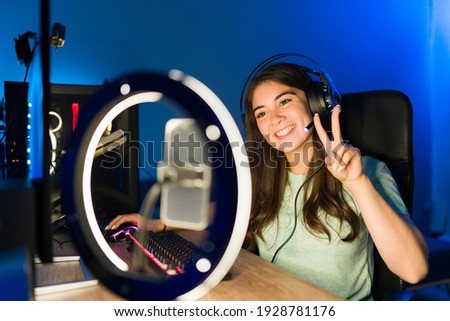 Portrait of a beautiful young woman saying hello to her online viewers during a live stream with a smartphone and a ring light. Smiling female gamer playing a video game in a gaming PC Royalty-Free Stock Photo #1928781176