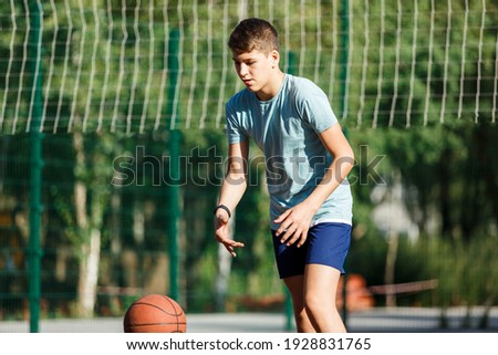 Cute Teenager in green t-shirt with orange basketball ball plays basketball on street playground in summer. Hobby, active lifestyle, sports activity for kids.	