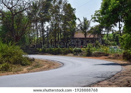 Picture of curved empty asphalt road shining in afternoon bright sunlight covered with green trees. small farmhouse constructed using clay made roof tiles beside the road at Kolhapur city India.