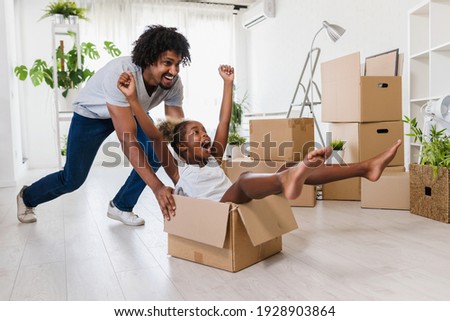 Father push cute little daughter sitting inside of carton box having fun riding in living room. Loan mortgage, housing improvement concept. Cheerful happy african family enjoy relocation day. Royalty-Free Stock Photo #1928903864