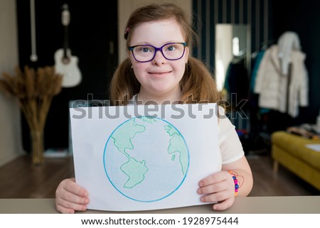 Portrait of teenage smiling girl with Down syndrome holding drawing of the planet Earth at home. Disabled child showing creative artwork. World Down syndrome day.