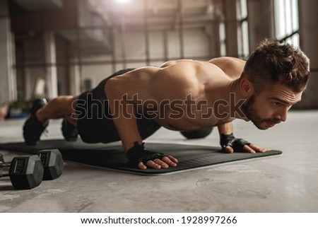Muscular man doing push up exercise on mat near dumbbells during intense fitness workout in grungy gym Royalty-Free Stock Photo #1928997266