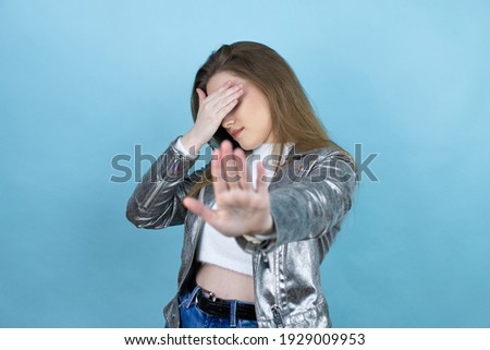 Pretty woman with long hair wearing a casual jacket over blue background covering eyes with hands and doing stop gesture with sad and fear expression. Embarrassed and negative concept.