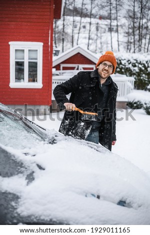 grey   electric car with Scandinavian house in the background. A nerdy guy with brush is removing snow.