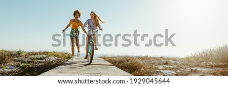 Excited woman riding bike down the boardwalk with her friends running along. Two female friends having a great time on their vacation. Panoramic shot with lots of copy space on background. Royalty-Free Stock Photo #1929045644