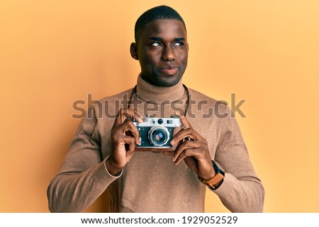 Young african american man holding vintage camera smiling looking to the side and staring away thinking. 