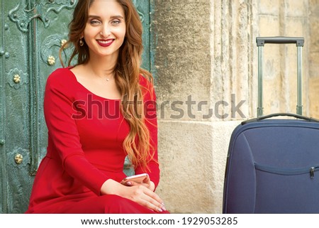 Portrait of a beautiful young caucasian traveling woman sitting at the door with suitcase smiling and looking at the camera outdoors