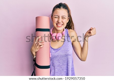 Young brunette woman holding yoga mat screaming proud, celebrating victory and success very excited with raised arm 