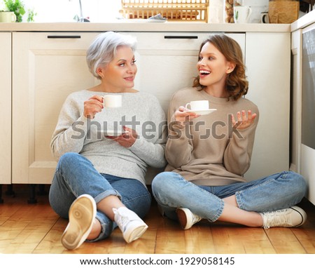 Cheerful young woman telling funny story to mature mother wearing casual clothes while sitting together on floor and drinking coffee during free time together at home Royalty-Free Stock Photo #1929058145