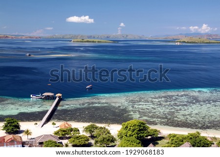 View of the sea strait, sandy beach with green trees, wooden pier. Coral reef and deep blue water.