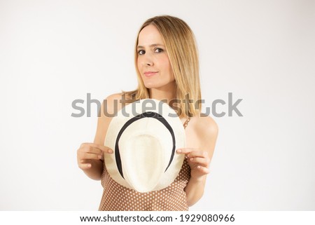 Pleased elegant woman in dress and straw hat posing while looking at camera over white background