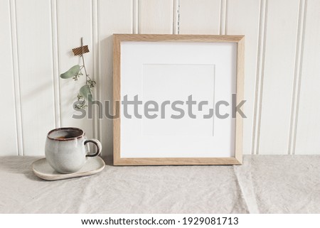Empty square wooden picture frame mockup. Cup of coffee on linen table cloth. Dry eucalyptus taped on  white wooden wall background. Home office concept. Scandinavian interior design.