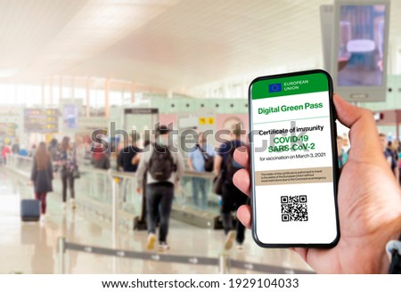 The digital green pass of the european union with the QR code on the screen of a mobile held by a hand with blurred airport in the background. Immunity from Covid-19. Travel without restrictions. Royalty-Free Stock Photo #1929104033