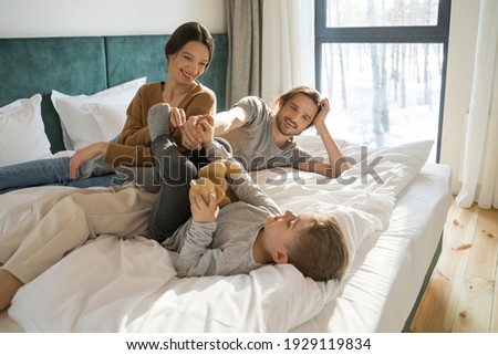 Family fun. Happy positive boy laying at the bed and holding his teddy bear while his parents looking at him with tenderness. Family relationships concept. Stock photo Royalty-Free Stock Photo #1929119834