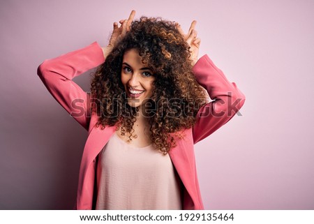 Young beautiful businesswoman with curly hair and piercing wearing elegant jacket Posing funny and crazy with fingers on head as bunny ears, smiling cheerful