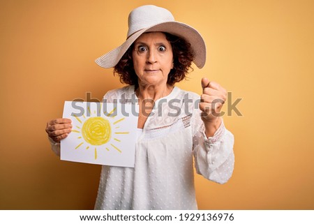 Middle age curly woman on vacation holding bunner with sun image over yellow background annoyed and frustrated shouting with anger, crazy and yelling with raised hand, anger concept
