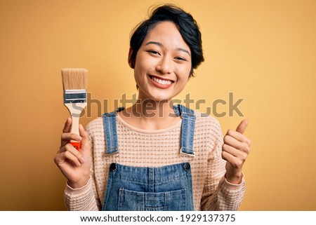Young beautiful chinese woman painting holding paint brush over isolated yellow background happy with big smile doing ok sign, thumb up with fingers, excellent sign