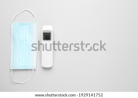 Infrared thermometer and medical mask on grey background, flat lay with space for text. Checking temperature during Covid-19 pandemic