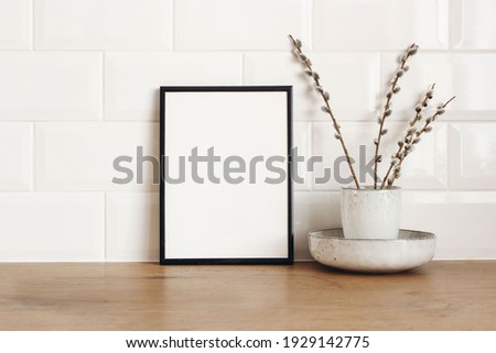 Spring still life. Blank black picture frame mockup on wooden table background. Easter composition with blooming goat willow, pussy willow or great sallow in ceramic vase. White tiles wall background.