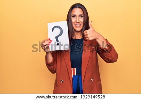 Young beautiful brunette woman holding paper with question mark smiling happy and positive, thumb up doing excellent and approval sign 