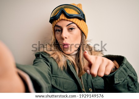 Young brunette skier woman wearing snow clothes and ski goggles make selfie by camera pointing with finger to the camera and to you, hand sign, positive and confident gesture from the front