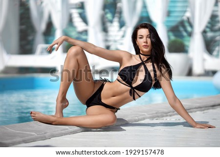 Beautiful brunette girl with perfect legs in the black bikini posing near the blue water of the swimming pool in the rays of sunset.