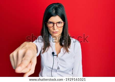 Young hispanic woman using lavalier microphone pointing with finger to the camera and to you, confident gesture looking serious 