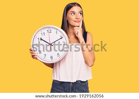 Young hispanic woman holding big clock serious face thinking about question with hand on chin, thoughtful about confusing idea  Royalty-Free Stock Photo #1929162056