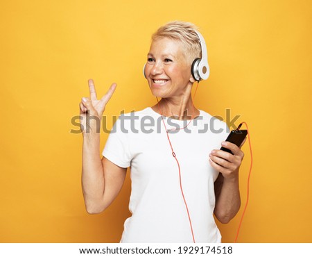 woman with short white hair wearing white tshirt listening to music with headphones and show victory sign over yellow background