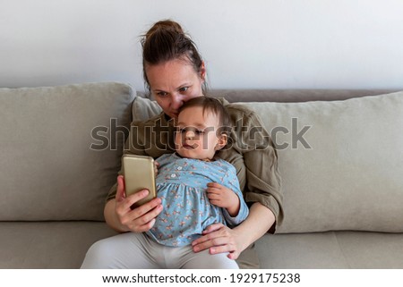 Mother and baby playing with a smartphone sitting on a sofa in the living room at home. Mom and baby girl having fun at home. Happy little daughter sitting on mother lap, family free time on internet.